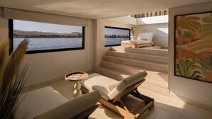 Una habitación moderna con un ventanal grande que da a una masa de agua, una tumbona, una mesita y una escalera que conduce a un nivel superior.
