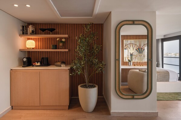 A modern hotel room with a wooden panel wall, a large mirror, a potted plant, a desk with a coffee maker, and a view of the city through the window. - Soleil Nile Cruise - Mondays from Luxor & Fridays from Aswan (Luxor)