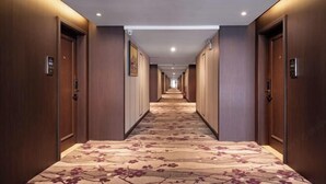 Un largo y estrecho pasillo de hotel con puertas de madera a ambos lados, una alfombra estampada e iluminación empotrada en el techo.