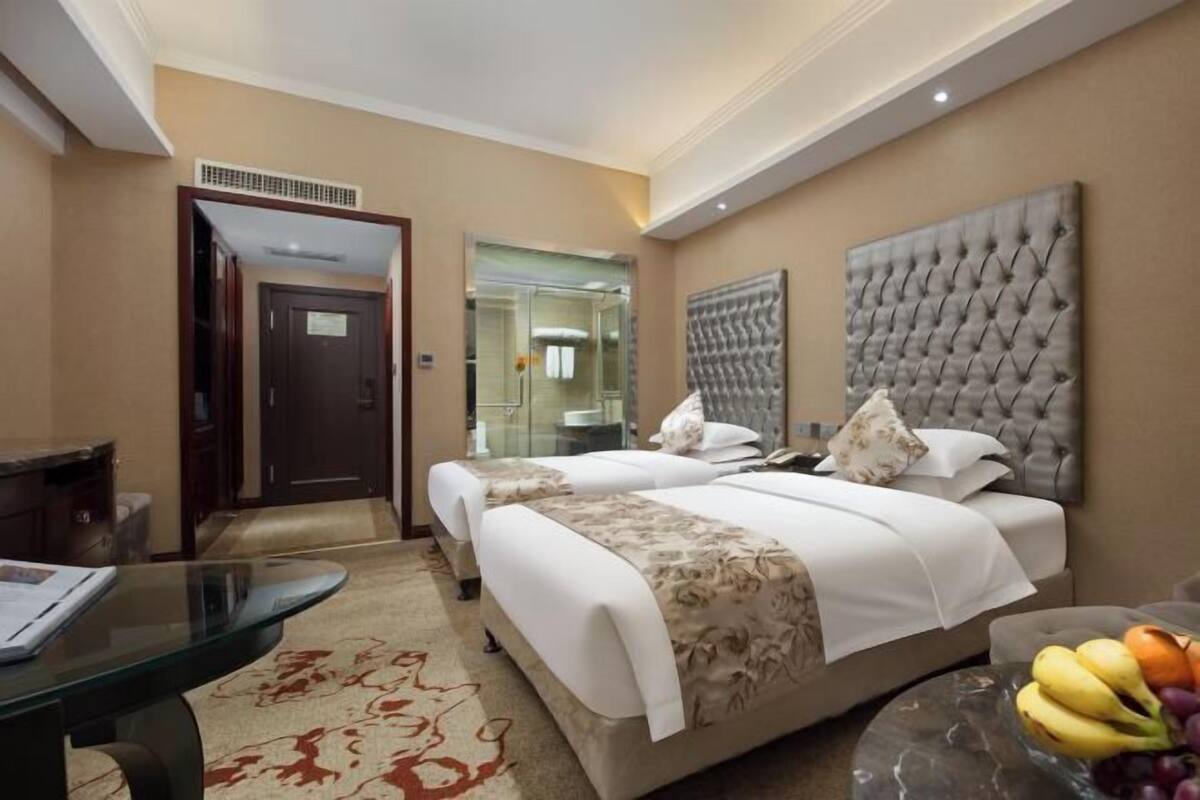 A hotel room with two beds, a glass table, a fruit bowl, and a large tufted headboard.