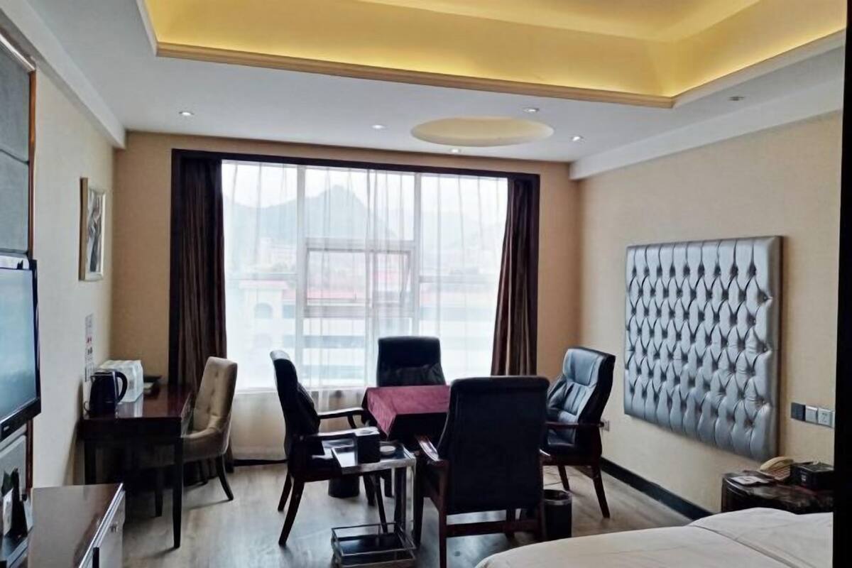 A hotel room with a bed, a dining table with chairs, a television, and a large window with curtains.