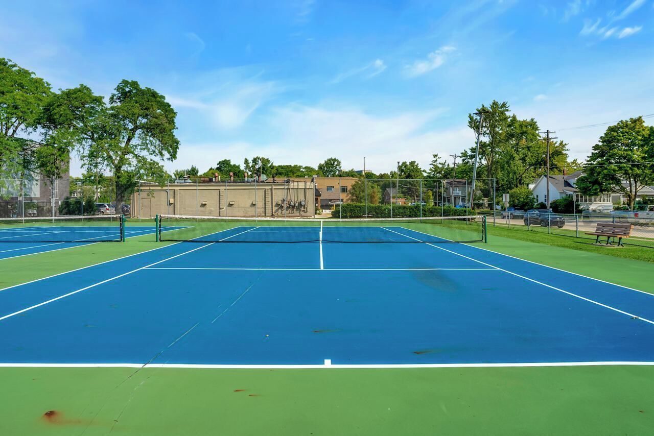 Un court de tennis en plein air, doté d’un filet, entouré d’arbres et de bâtiments.
