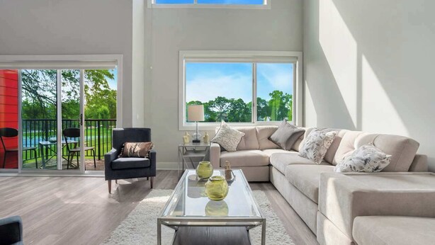 A spacious living room with a large glass coffee table, a beige sofa, and a view of a green outdoor area through large windows.