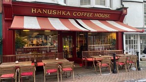 Ein Kaffeehaus mit roten und weißen Streifen an der Markise, Holztischen und -stühlen, und einem Schild mit der Aufschrift "HANUSHKA COFFEE HOUSE".