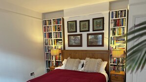 Ein ordentlich bezogenes Bett mit einer roten Decke und weißen Kissen, eingerahmt von zwei Nachttischen mit Lampen. Dahinter ist eine Bücherwand mit Büchern zu sehen.