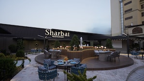 Un espace de restauration en plein air avec des tables et des chaises rondes, un bar central et une enseigne indiquant « Restaurant Sharbat ».