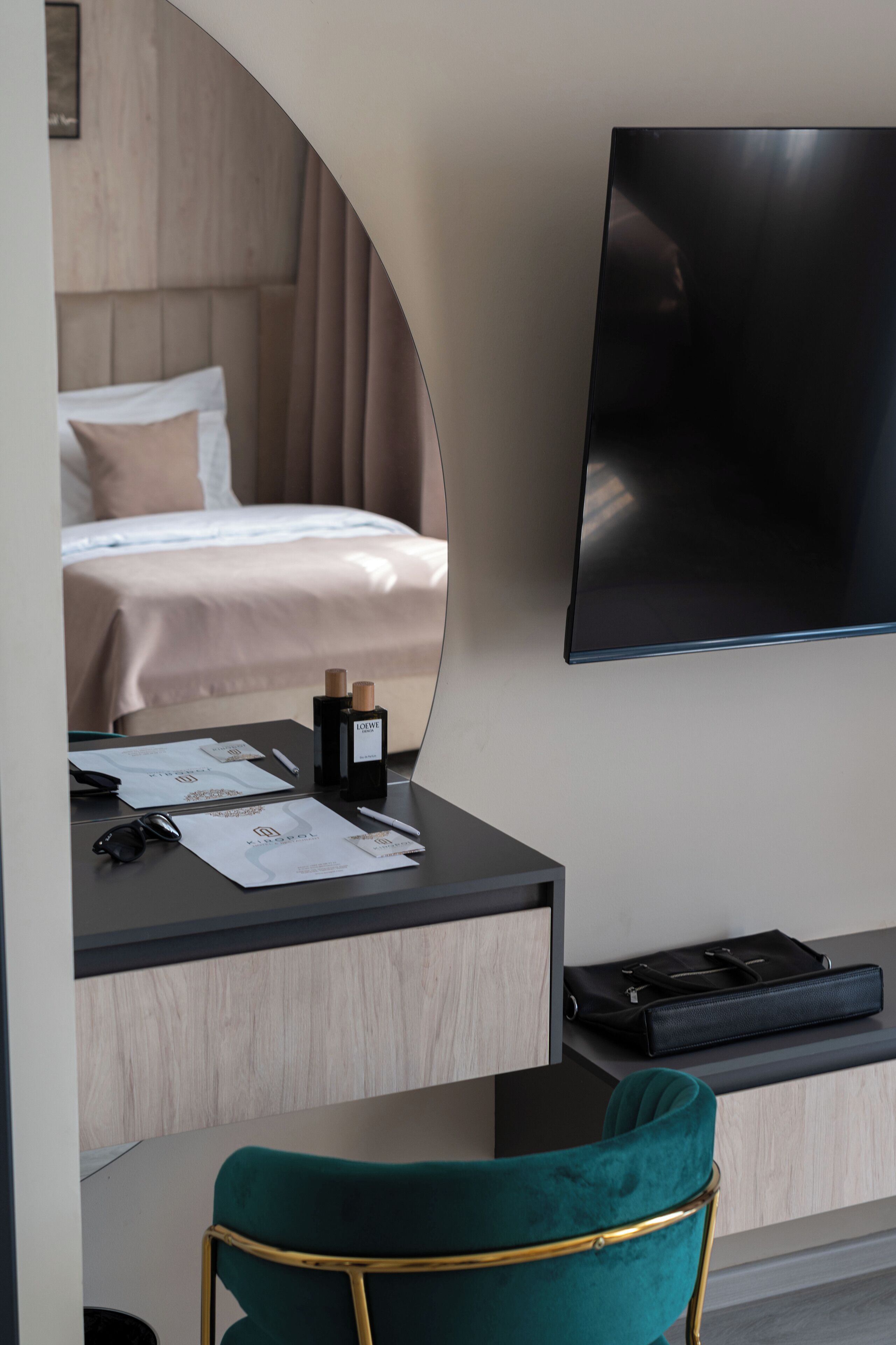 A modern hotel room with a bed, a desk with a mirror, a television, and a teal chair.
