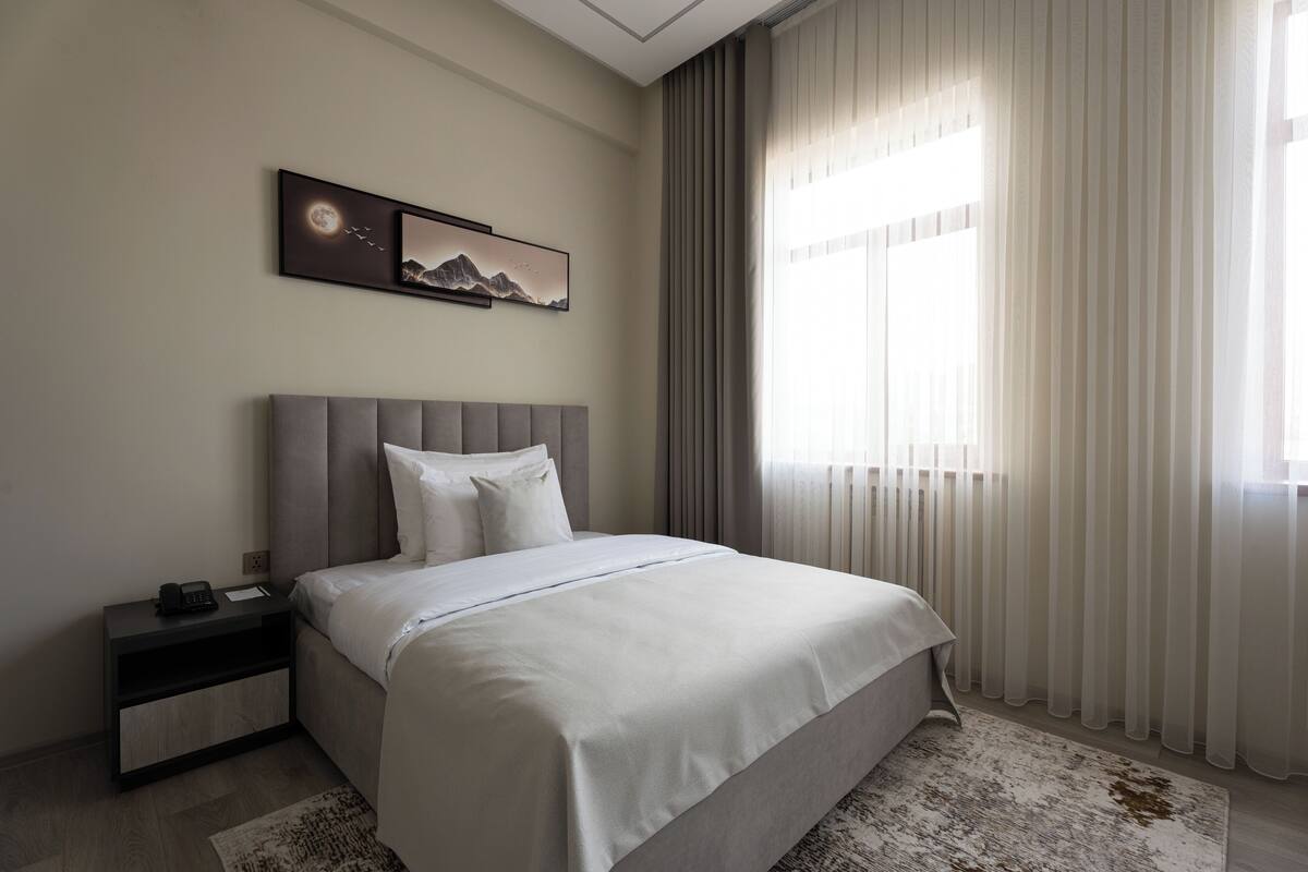 Ein modernes Hotelzimmer mit einem großen Bett, einem Nachttisch mit Telefon und einem Fenster mit durchscheinenden Vorhängen.
