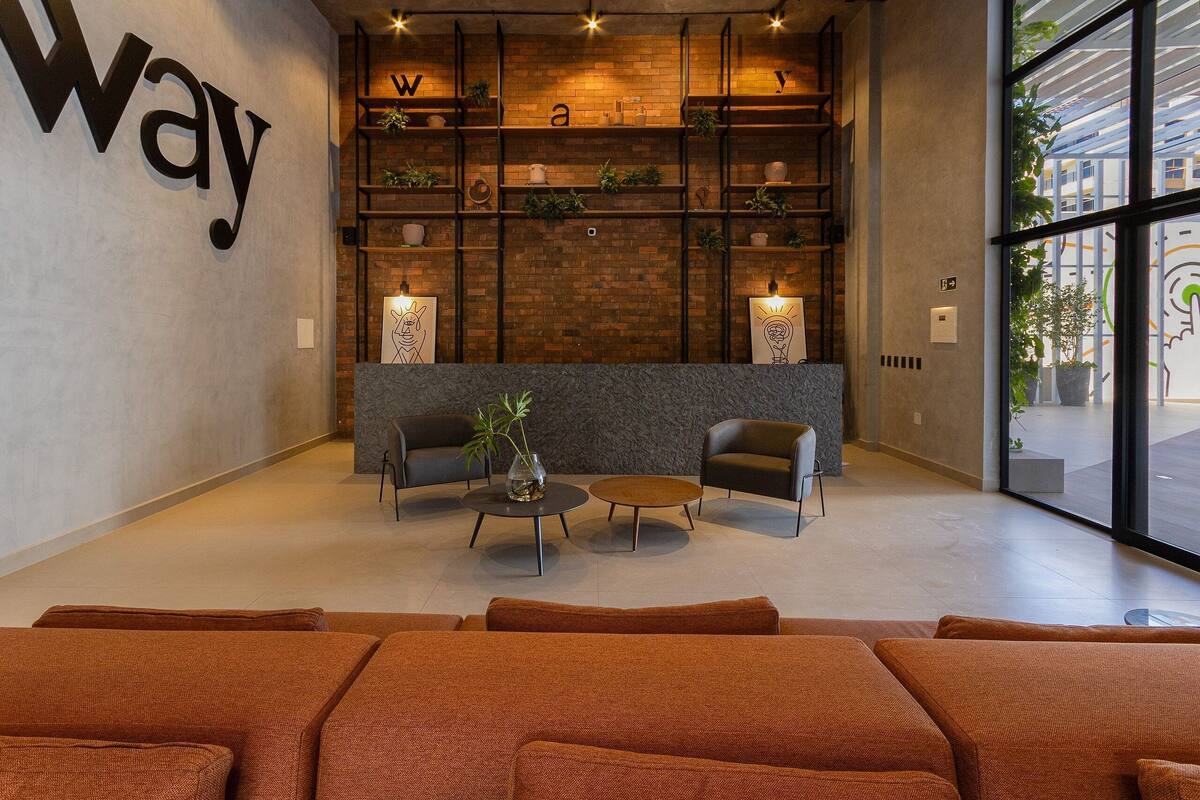 A modern lobby with a large 'way' sign, a brick wall, shelves with plants, two armchairs, a coffee table, and a large window.