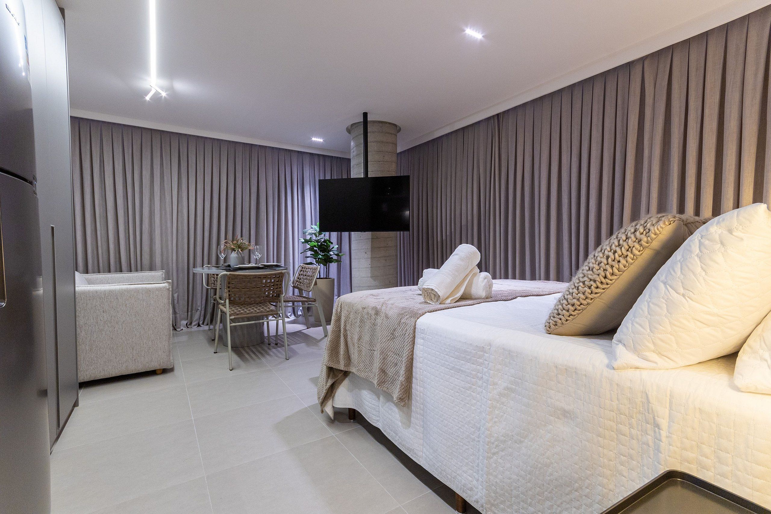 A modern hotel room with a large bed, a flat-screen TV mounted on the wall, a desk with a chair, and a small table with a plant.
