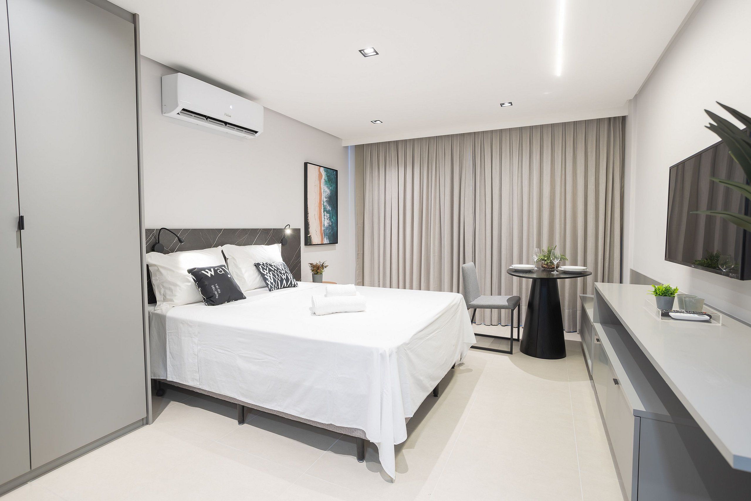 Ein modernes Hotelzimmer mit einem ordentlich bezogenen Bett, einem Esstisch mit Stühlen, einem kleinen Topfpflanzen und einem flachbildfernseher an der Wand.