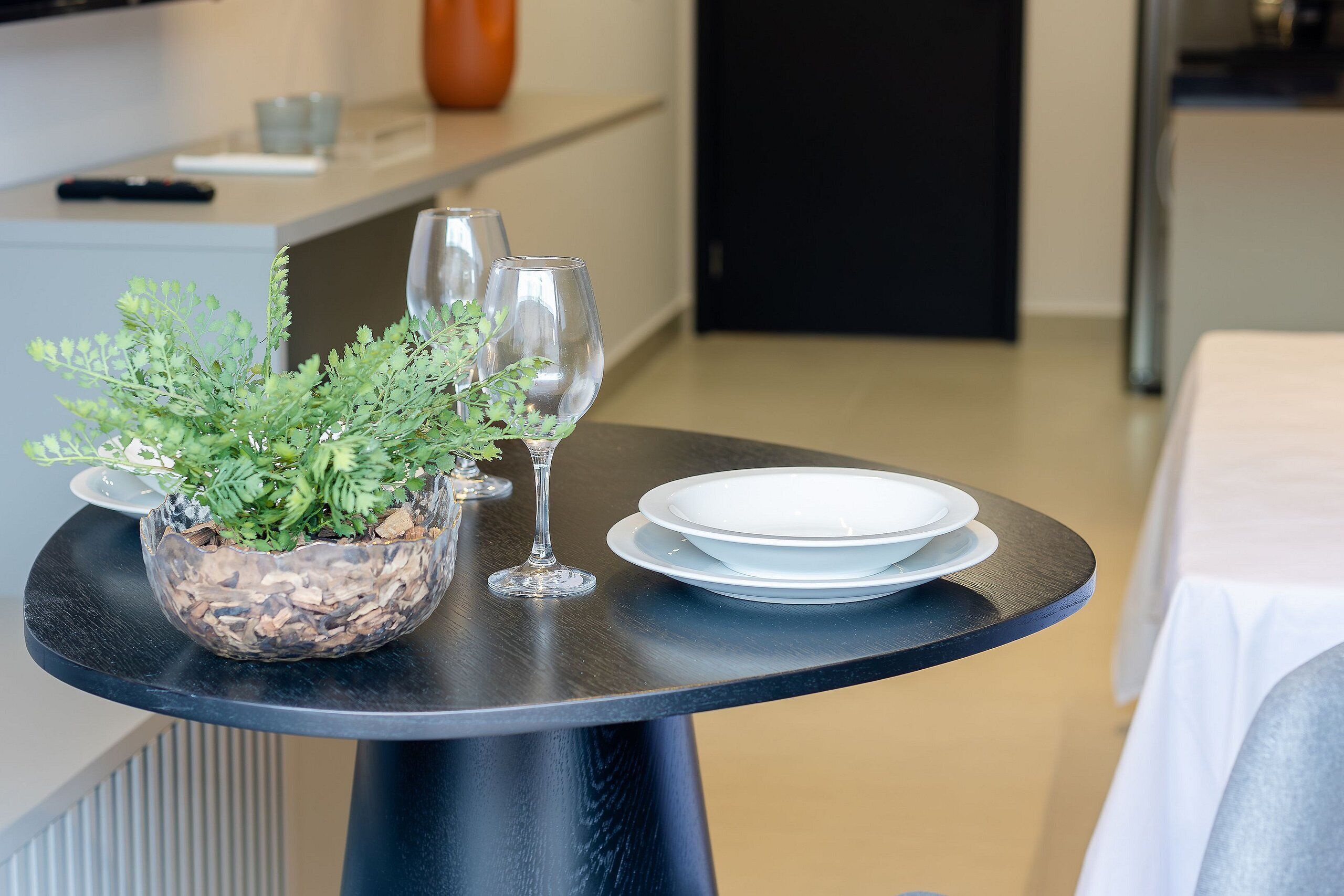 Una mesa de comedor con una planta en maceta, copas de vino y platos apilados en una habitación de hotel.