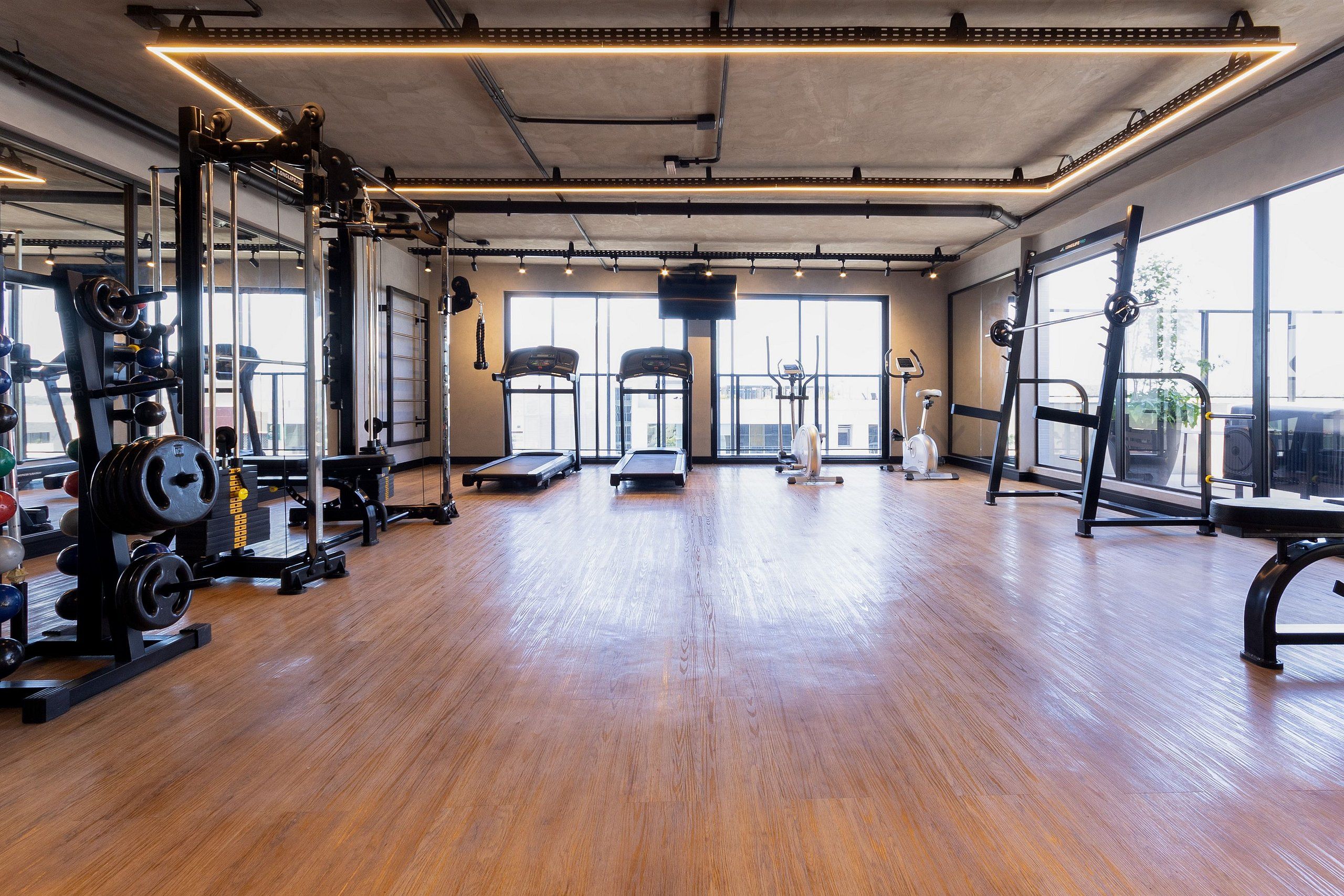 Een ruime fitnessruimte met houten vloer, diverse fitnessapparaten en grote ramen voor natuurlijk licht.