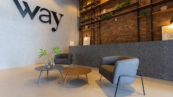 A modern lobby with a large 'way' sign, a brick wall with shelves, and two grey armchairs.
