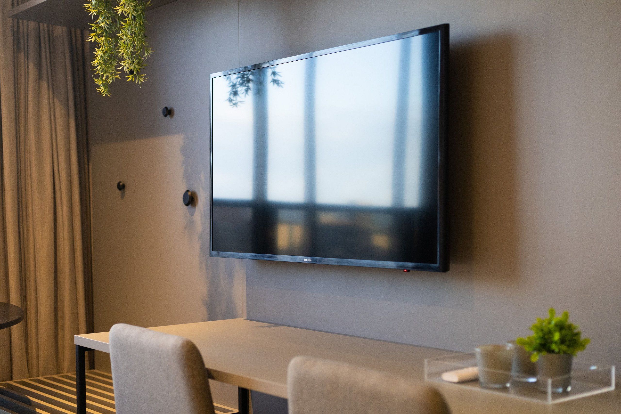Ett modernt rum med en platt-TV som är monterad på väggen, ett matbord med stolar, en liten krukväxt och ett fönster med gardiner.