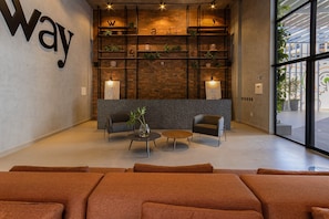 Um lobby moderno com um grande letreiro indicando 'way', uma parede de tijolos, prateleiras com plantas, duas poltronas, uma mesa de centro e uma janela ampla.
