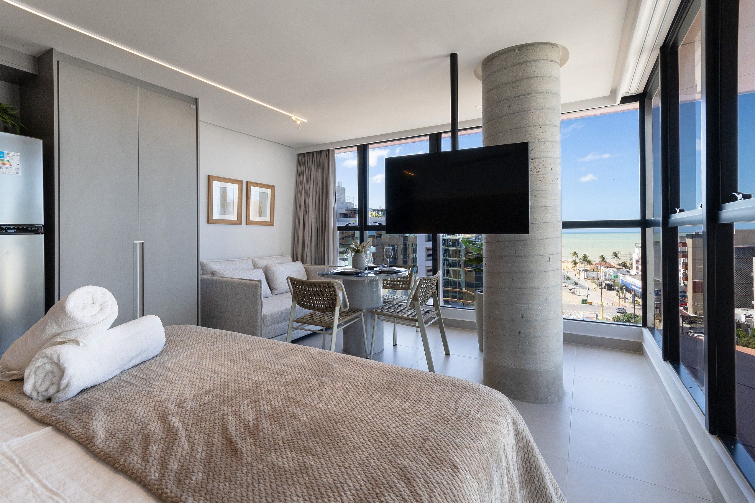 A modern hotel room with a large bed, a flat-screen TV, a balcony with a view of the beach, and a dining area with a table and chairs.