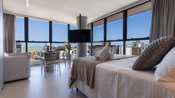 A modern hotel room with a large bed, a TV mounted on a pillar, a dining area with a table and chairs, and a view of the city and beach through large windows. - Way Tambaú - João Pessoa by Carpediem (João Pessoa)
