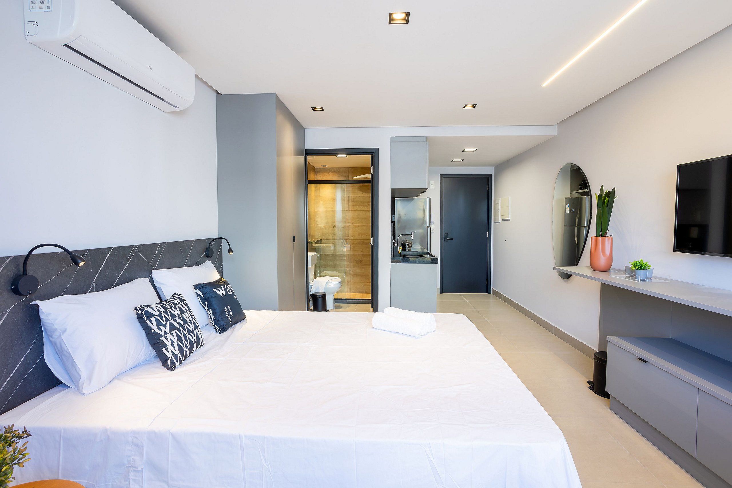 A modern hotel room with a large bed, a flat-screen TV, a small plant, and a bathroom visible through an open door.