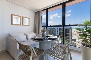 Un salon moderne avec un canapé, une table à manger et des chaises, de grandes fenêtres donnant sur la ville et une plante en pot.