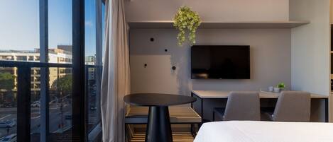 A modern hotel room with a large window, a round black table, a bed, a TV mounted on the wall, and a hanging plant.