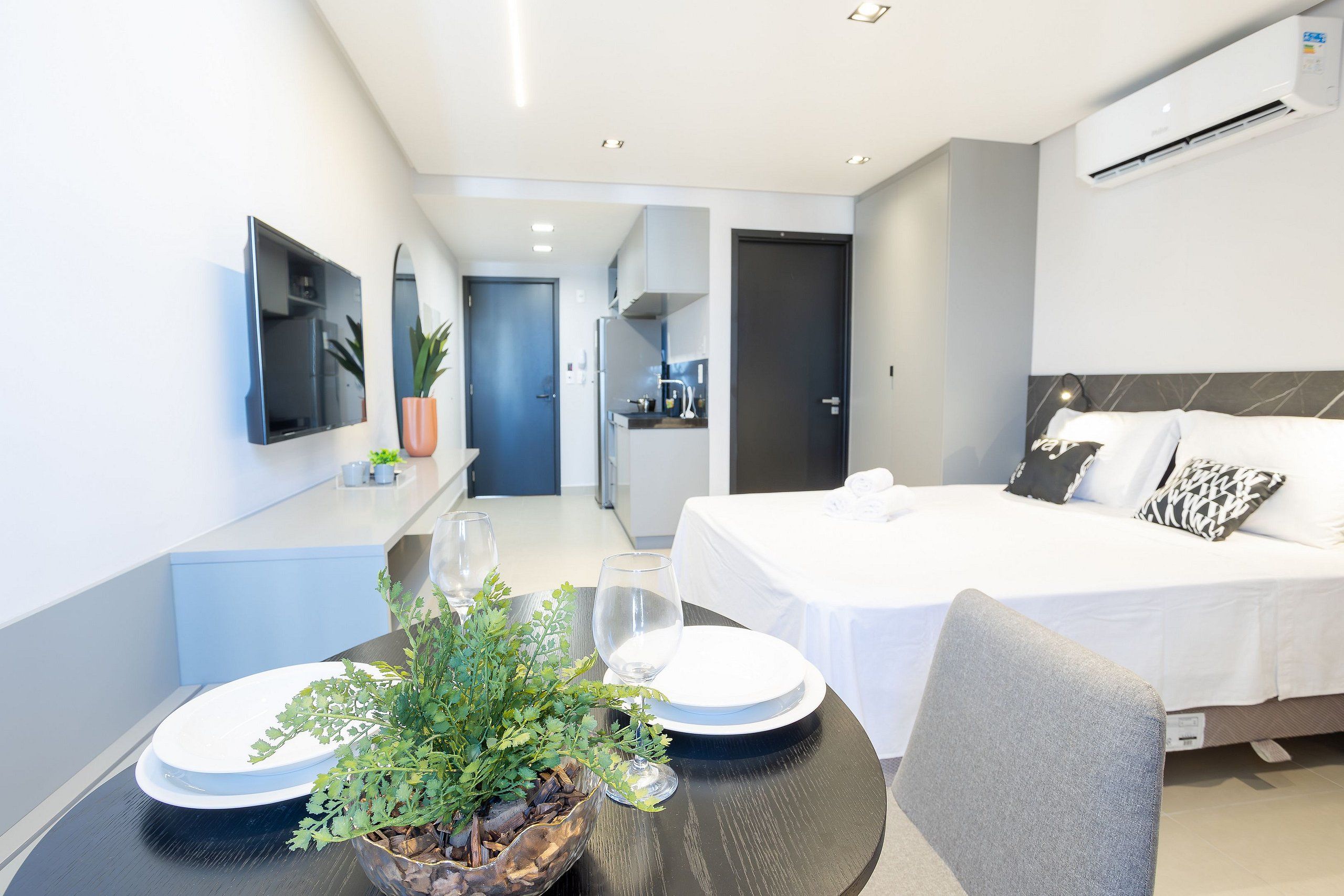 Et moderne hotelværelse med en seng, et spisebord med tallerkener og glas, en potteplante og et køkkenområde, der ses igennem en åben dør.