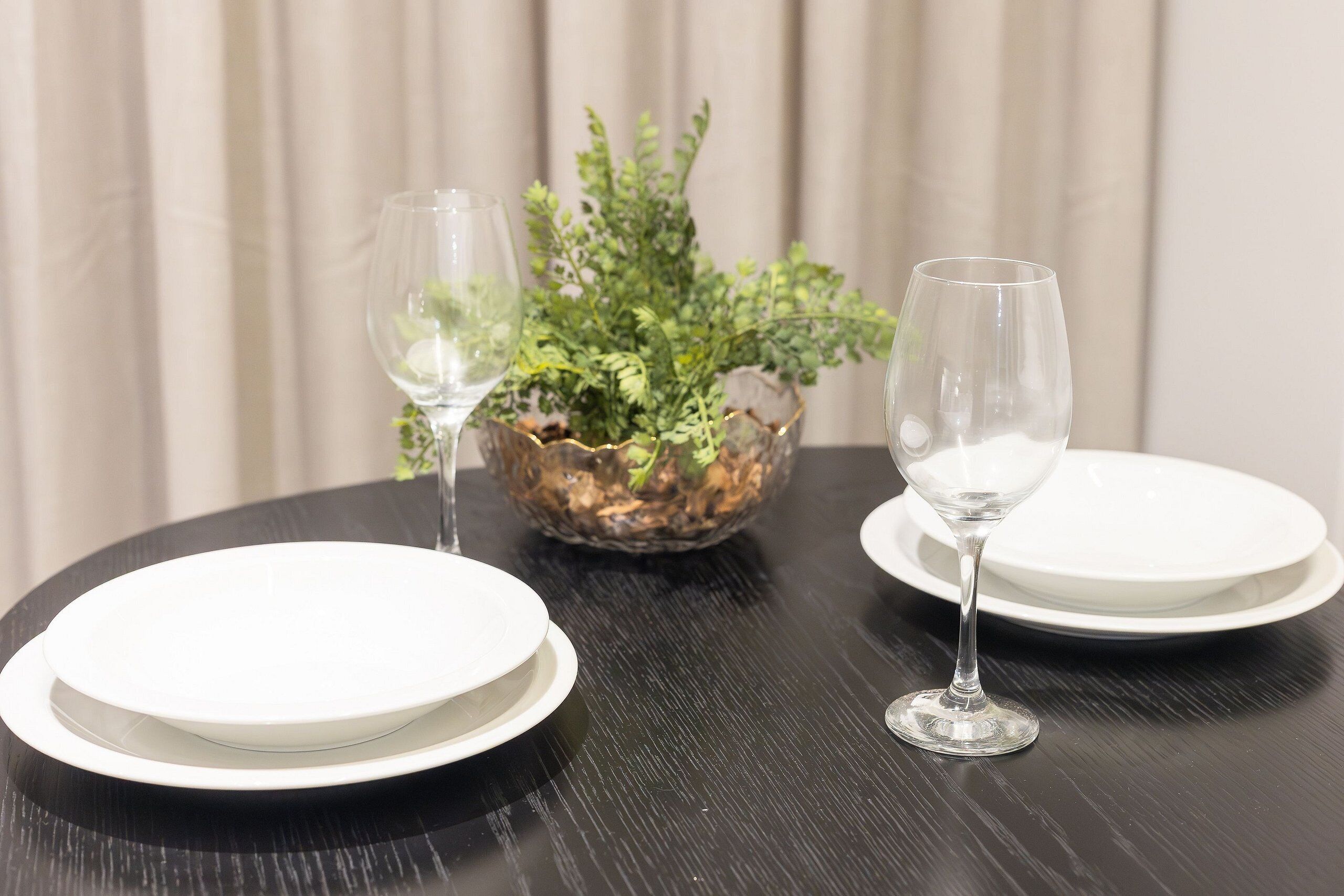 Een eettafel met twee witte borden en twee wijnglazen, een potplant en een gordijn op de achtergrond.