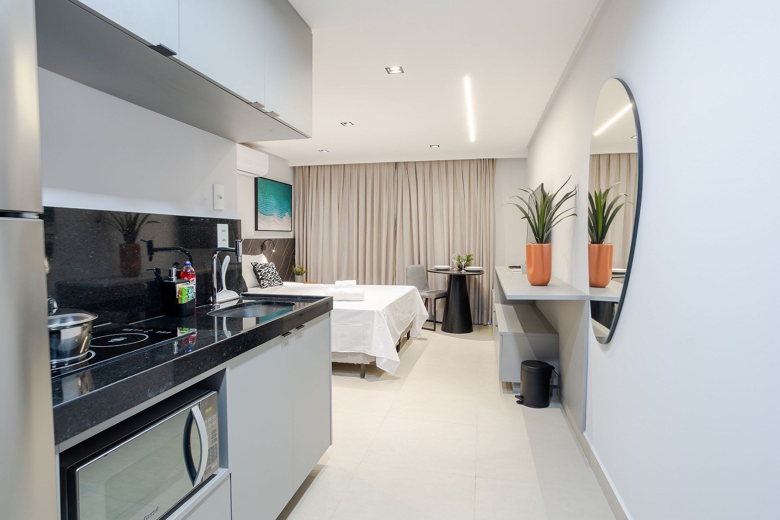 A modern kitchen with a black countertop, a microwave, and a stove. There is a dining area with a table and chairs, and a bed with a white bedspread.