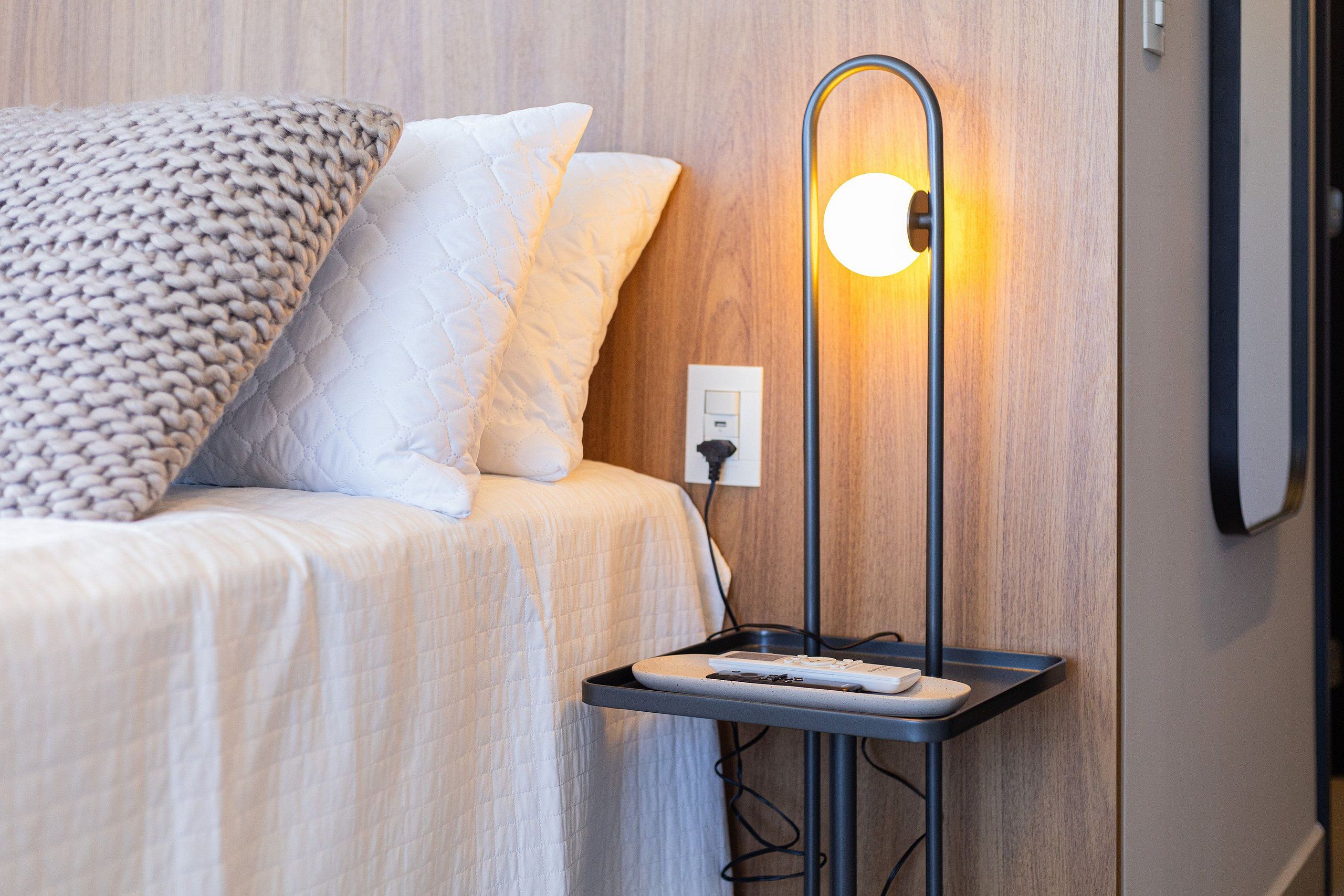 A neatly made bed with a white quilt and a knitted throw, a bedside table with a lamp, and a wall socket.