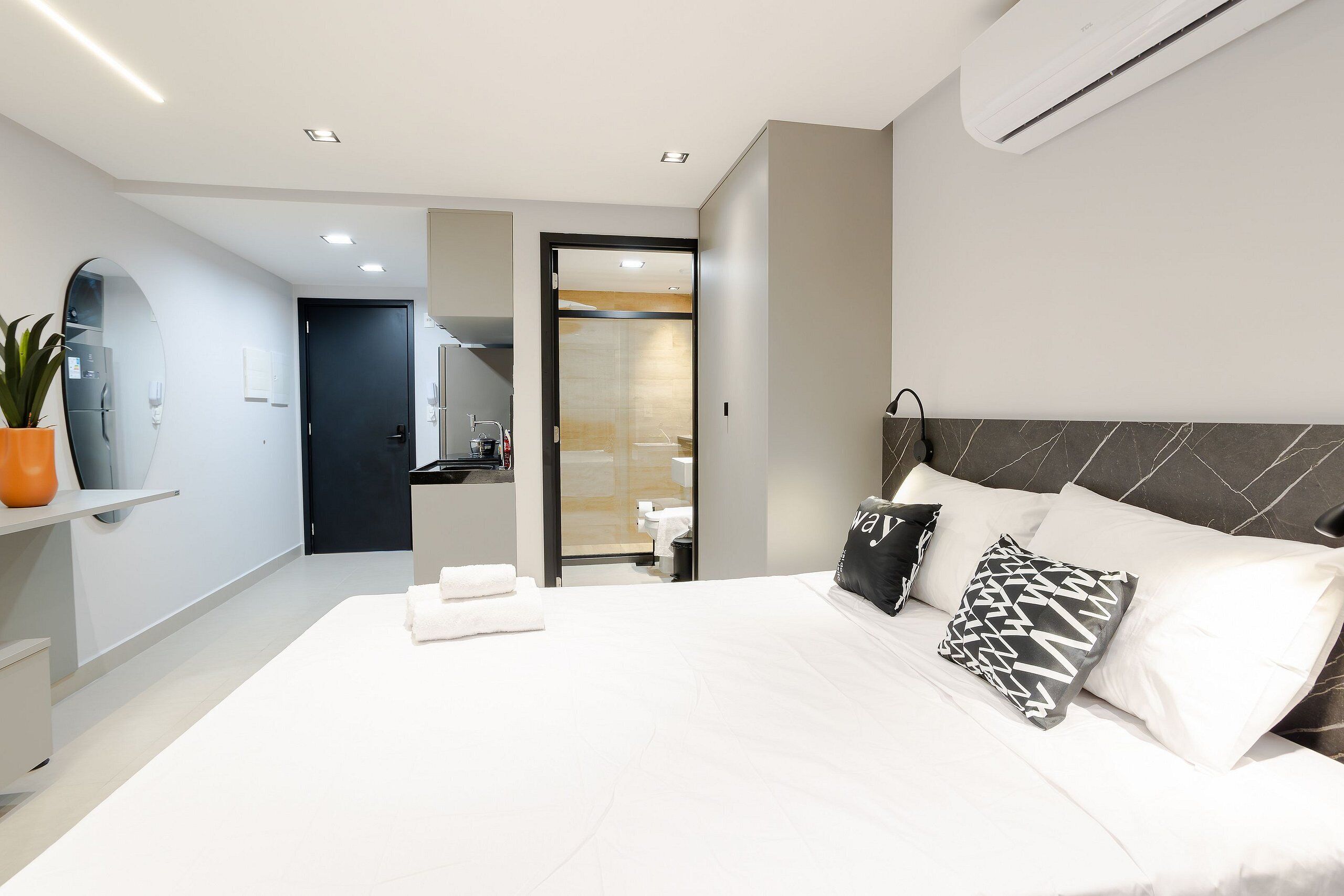 A modern bedroom with a large bed, a bedside table, a potted plant, and a bathroom visible through an open door.