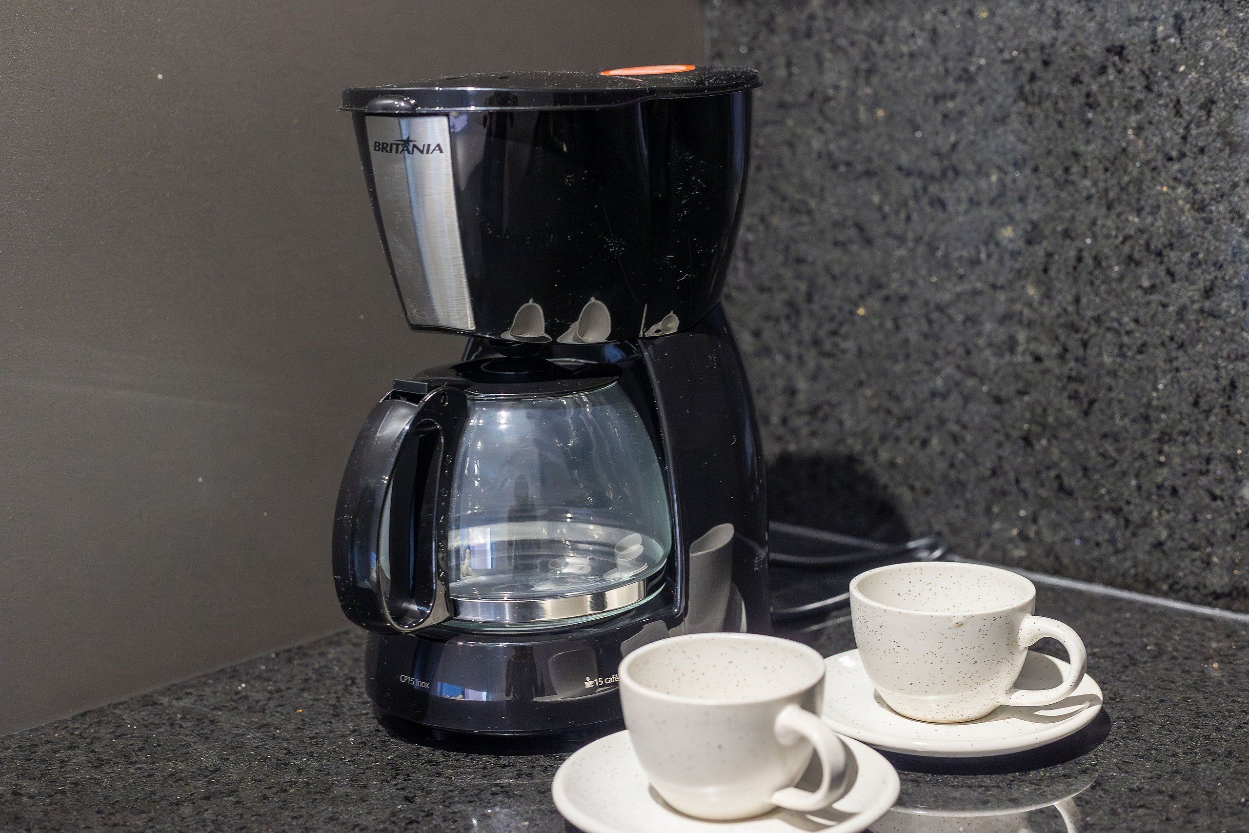 A black coffee maker with a glass carafe and two white cups on a granite countertop.