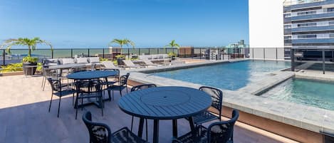 A rooftop pool area with lounge chairs, a round table, and a view of the ocean.