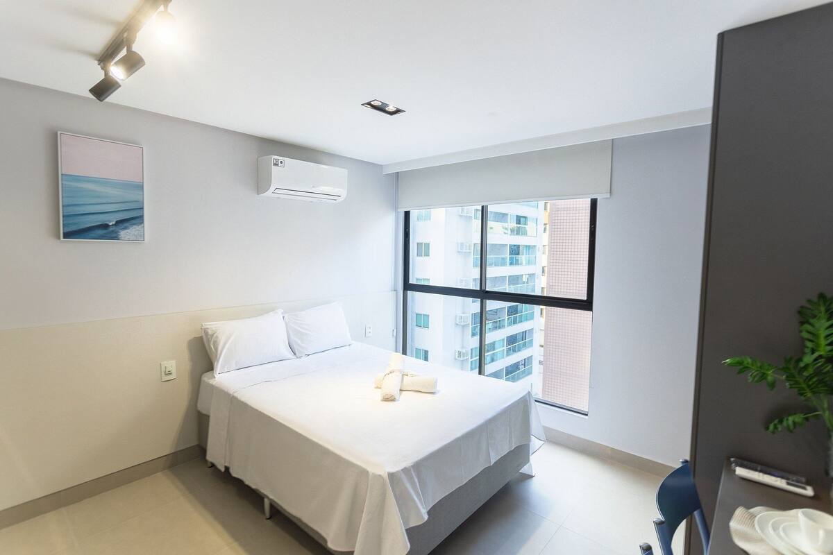 A modern hotel room with a bed, a window with a city view, an air conditioning unit, and a small table with a plant.