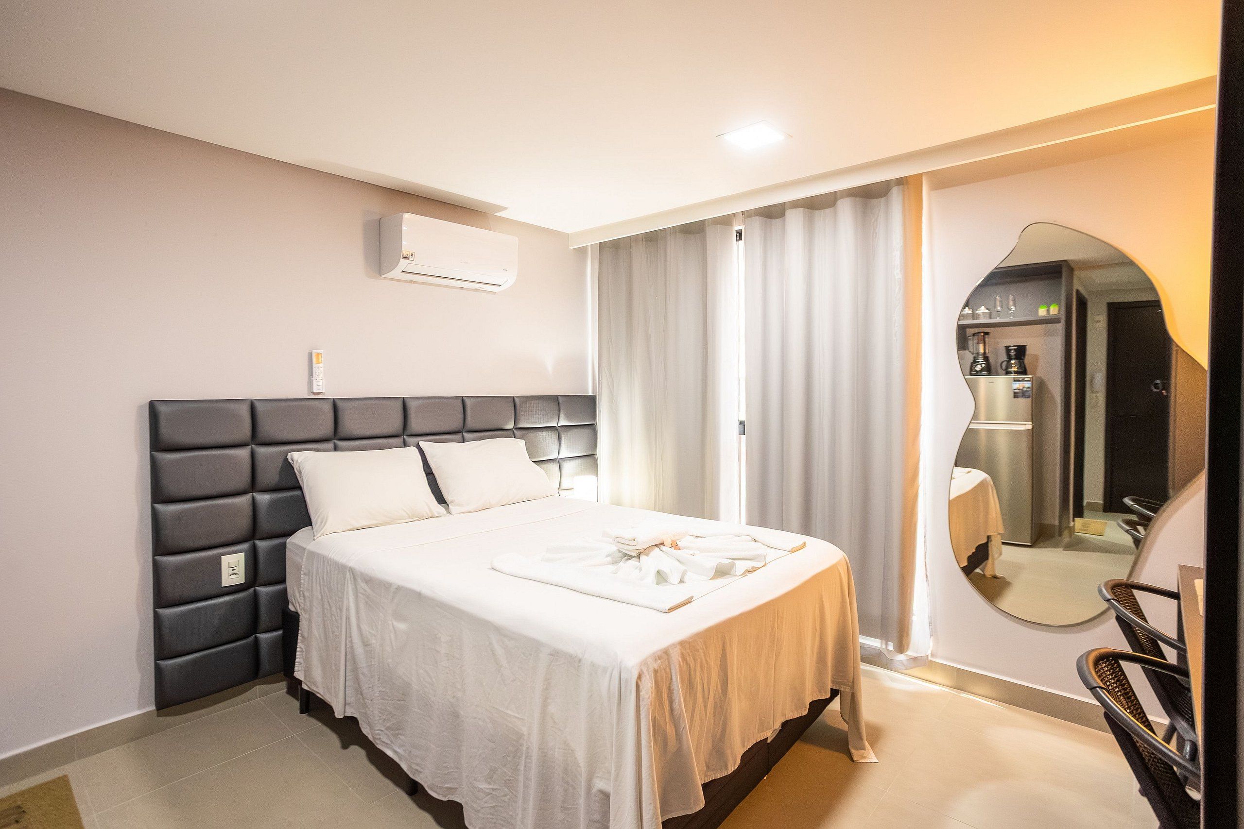 A hotel room with a bed, a bedside table, an air conditioning unit, a mirror, and a view of the bathroom.