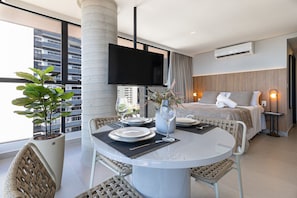 Quarto de hotel moderno com área de refeições, uma cama e uma janela grande com vista para a cidade.