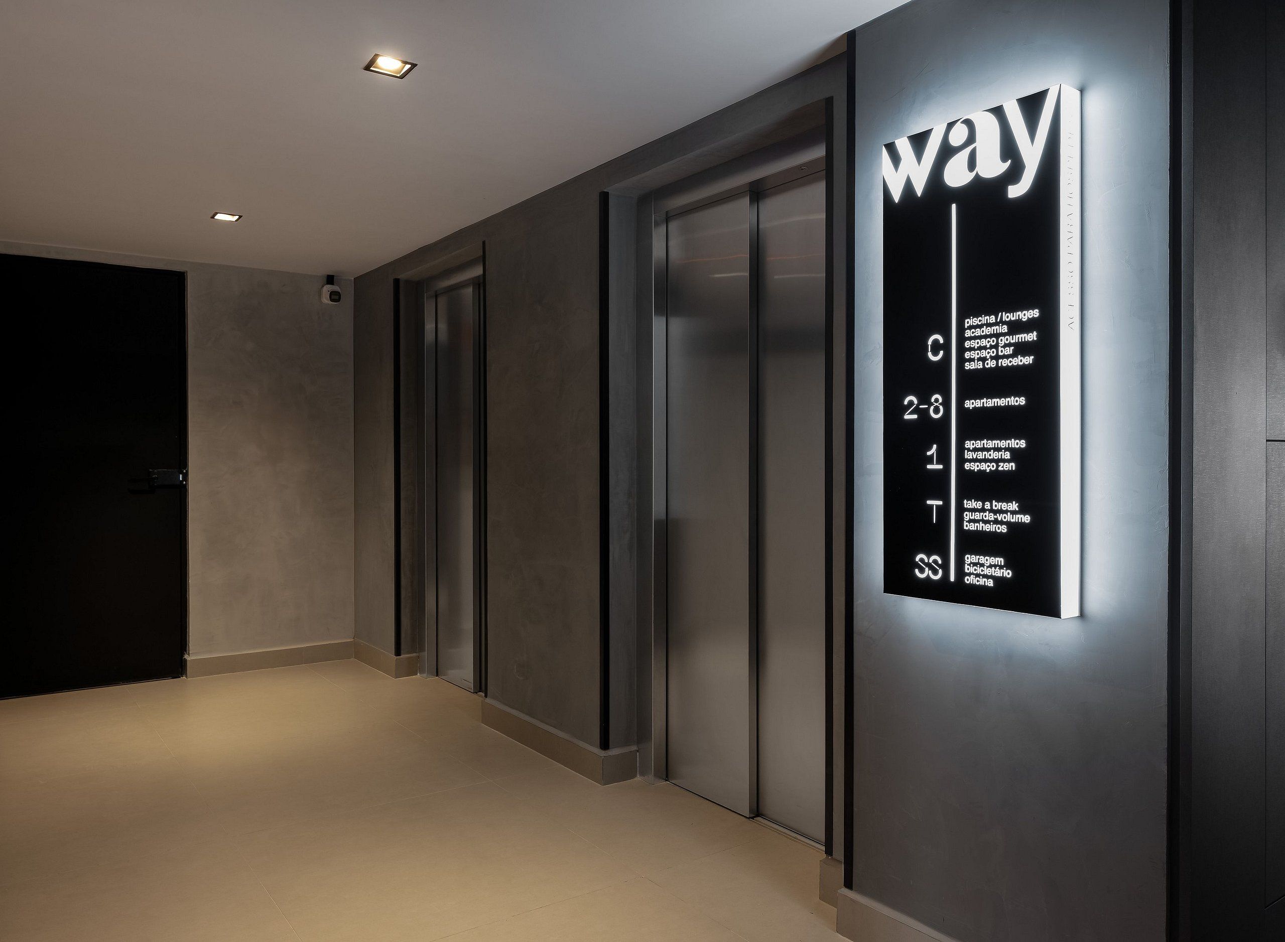 Un couloir doté d’un ascenseur moderne, d’un panneau mural indiquant les types de chambres et leurs numéros, et d’une porte au bout du couloir.