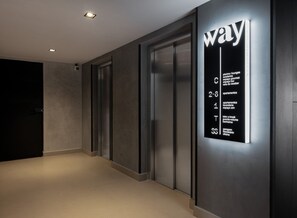A hallway with a modern elevator, a wall-mounted sign with room types and numbers, and a door at the end. - Way Tambaú - João Pessoa by Carpediem (João Pessoa)