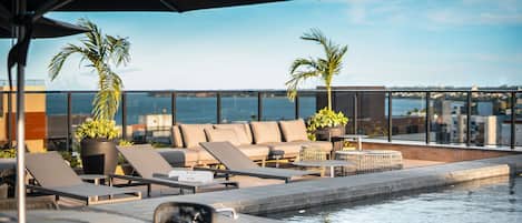 Un área de piscina en la azotea con sillones de descanso, una mesita y un gran paraguas.