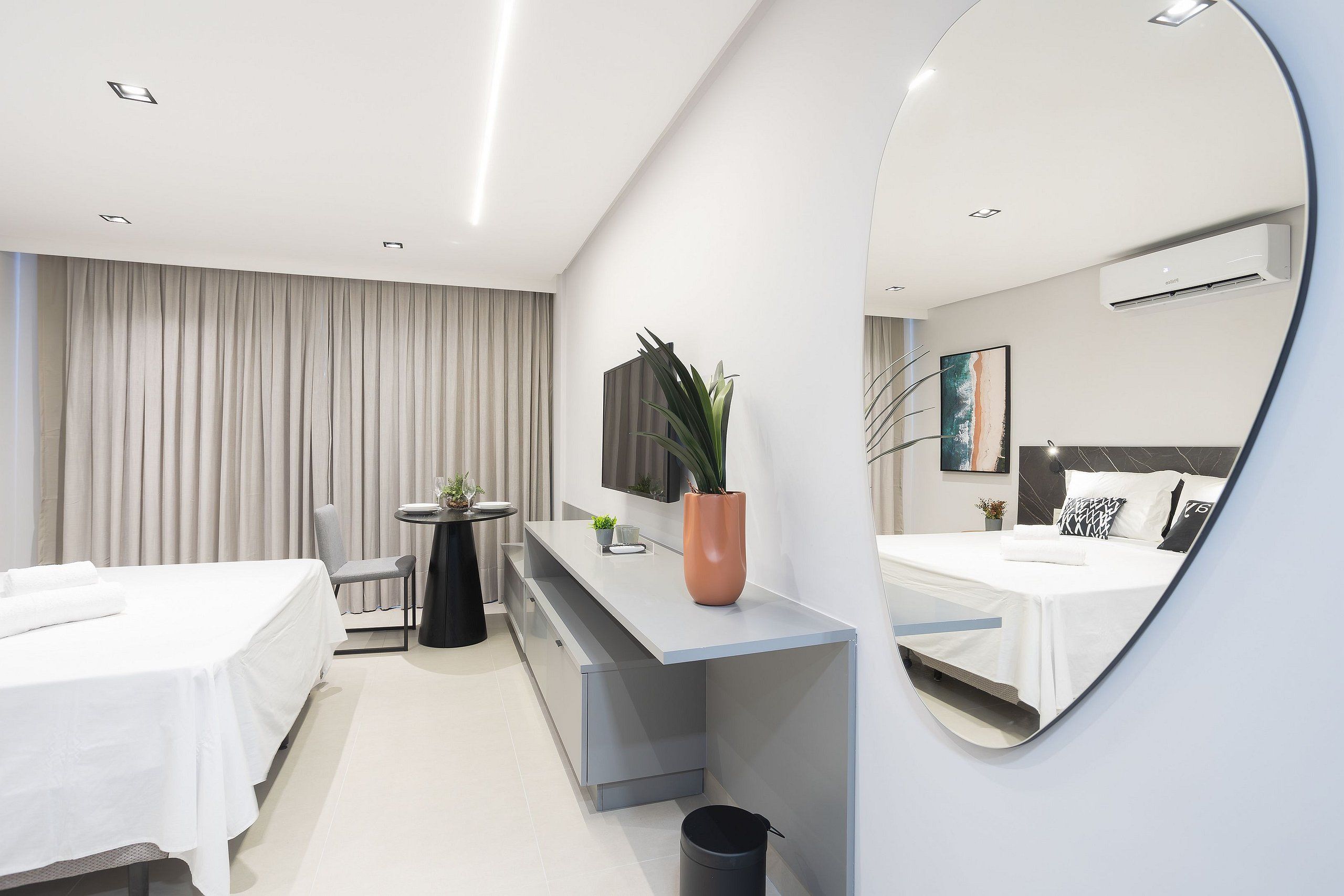 A modern hotel room with a bed, a desk, a chair, a potted plant, a small table, and a large mirror.
