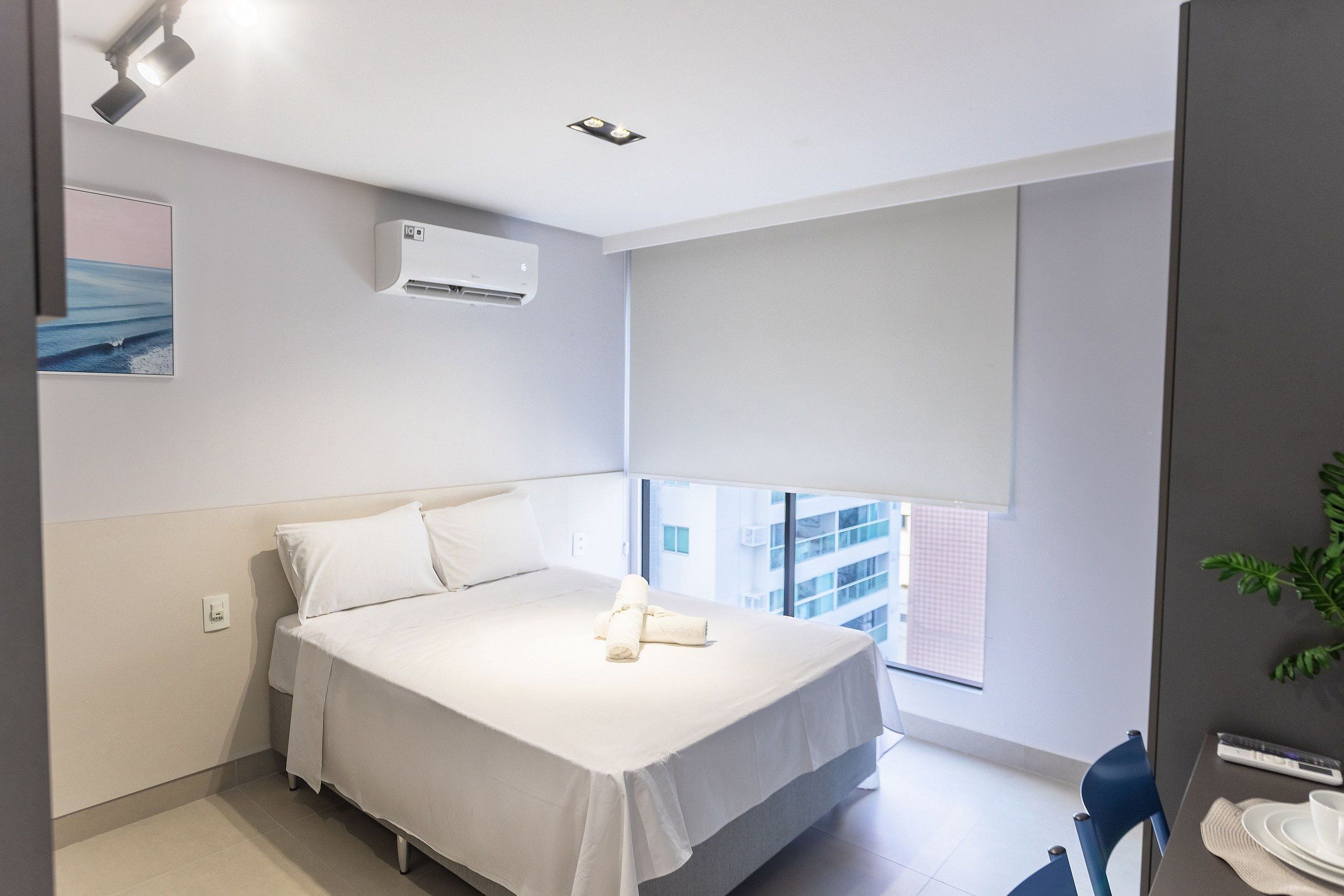 Habitación de hotel moderna con una cama grande, una ventana con persianas, un aire acondicionado instalado en la pared y una pequeña mesa con una silla.
