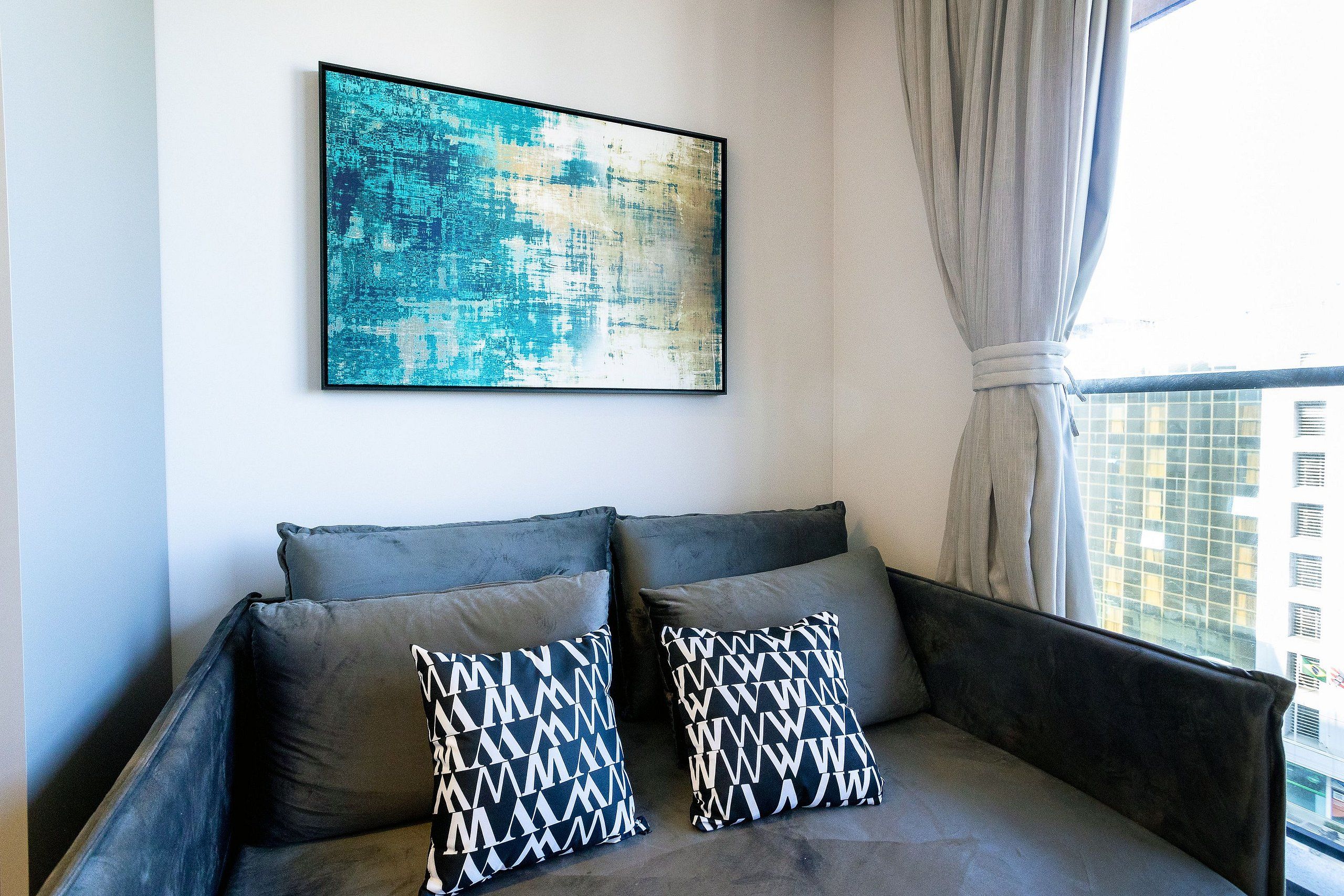 Ein modernes Wohnzimmer mit einem grauen Sofa, zwei gemusterten Kissen, einem großen abstrakten Gemälde und Blick auf eine Stadtlandschaft durch ein Fenster mit Vorhängen.