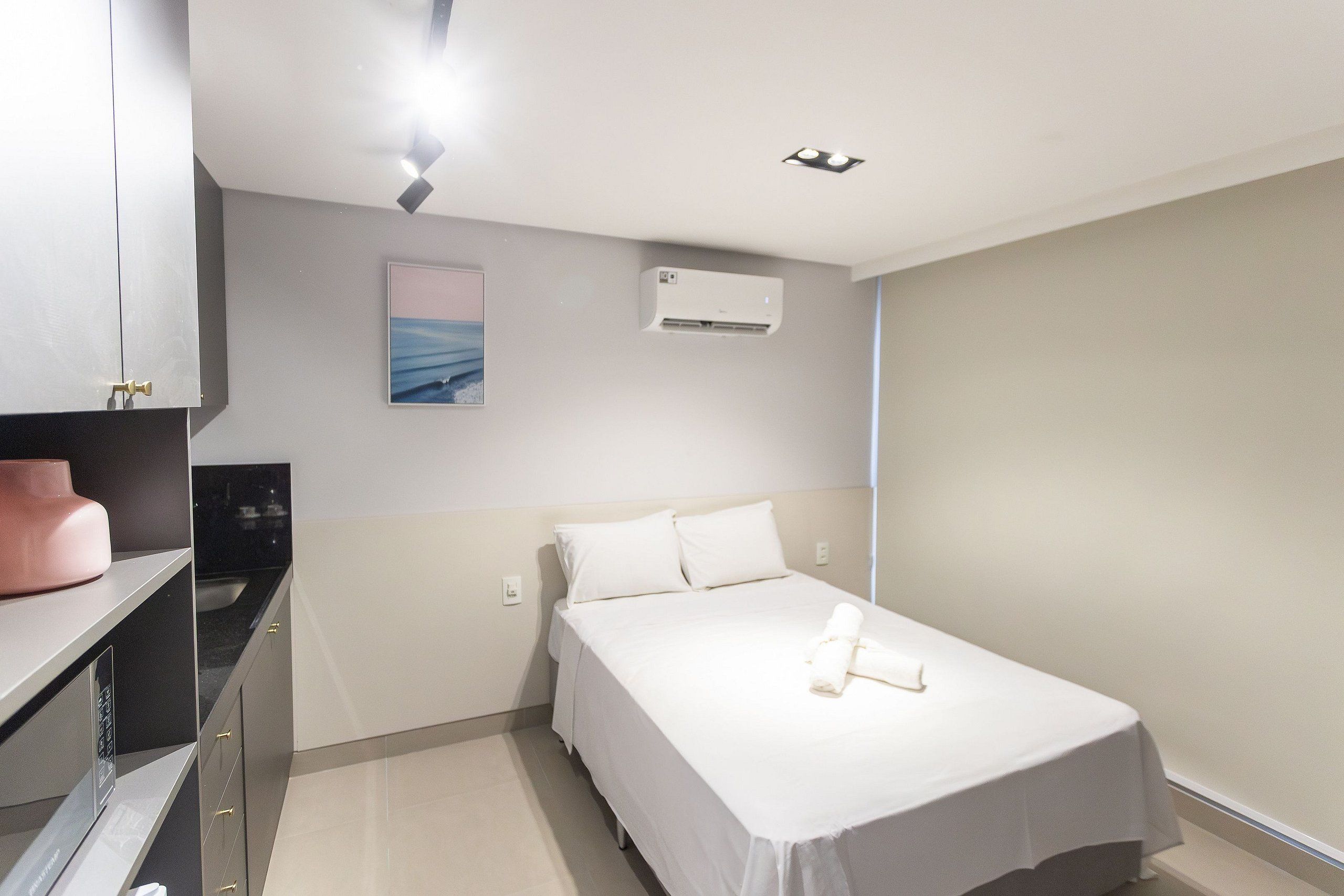 A hotel room with a bed, a small desk, a wall-mounted air conditioner, and a painting on the wall.