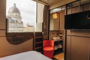 Une chambre d’hôtel avec une grande fenêtre, un fauteuil rouge, un téléviseur à écran plat et une étagère en bois.