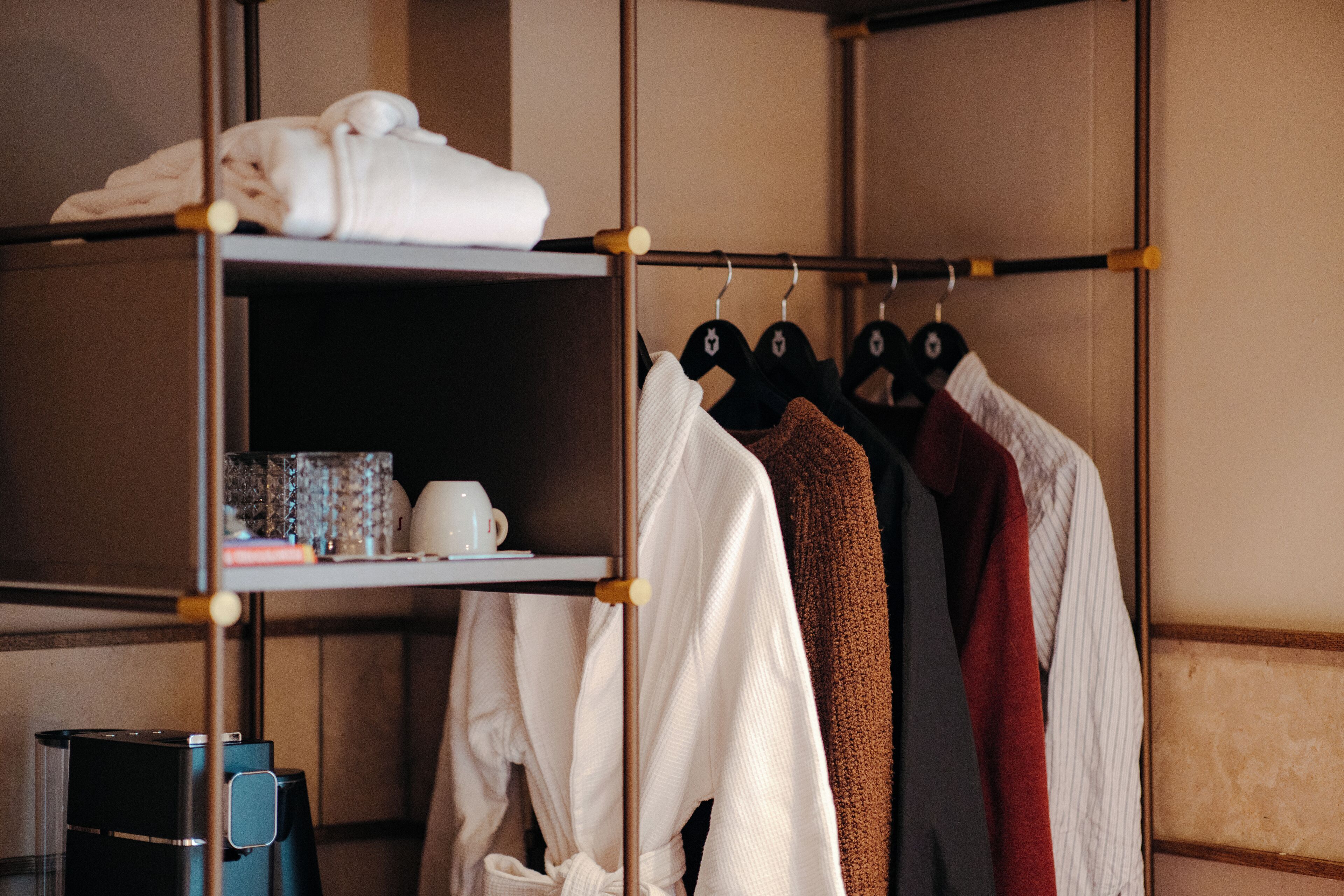 Een hotelkamer met een kledingkast vol hangende kleding en een plank met een handdoek en een beker.