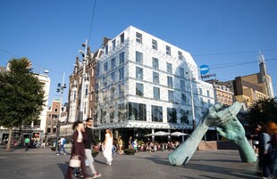 Un bâtiment moderne doté d’une façade géométrique originale, entouré d’une place animée où se croisent les piétons et où l’on trouve des espaces de restauration en plein air.