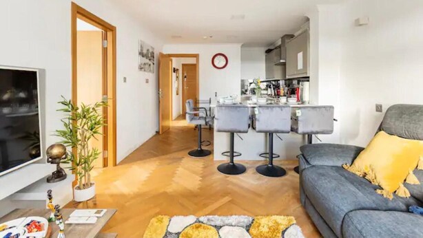 A modern living room with a grey sofa, a TV, and a dining area with bar stools. - 4 Guests - 2 Bedrooms - Quick Links Central London (London)
