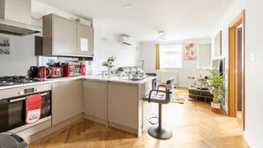 A modern kitchen with a central island, a stove, and a sink. There is a dining area with a bar stool and a plant. - 4 Guests - 2 Bedrooms - Quick Links Central London (London)