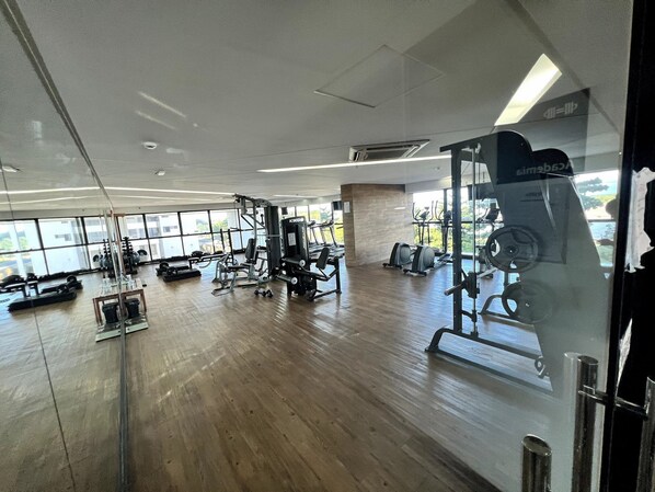 A spacious gym with various exercise equipment, large windows, and a wooden floor.