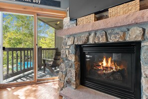 A stone fireplace with a glass screen, a pool visible through a sliding glass door, and a wooden deck with outdoor furniture. - Short Walk to Lifts & Village Amenities & Private Balcony Overlooks Pool! (Keystone)