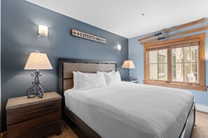 A bedroom with a large bed, two bedside tables with lamps, a wooden headboard, and a window with blinds.